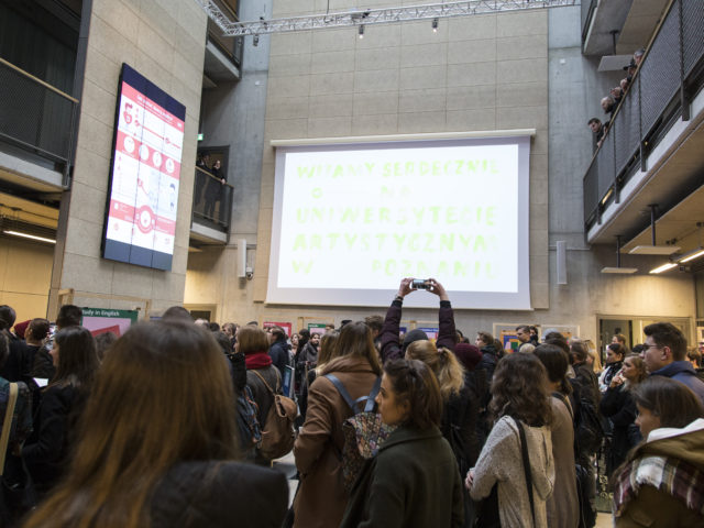 Ludzie zgromadzeni w sali Atrium w budynku B Uniwersytetu Artystycznego w Poznaniu podczas Drzwi Otwartych pomiędzy stoiskami poszczególnych kierunków i wydziałów; na rzutniku napis powitylany.
