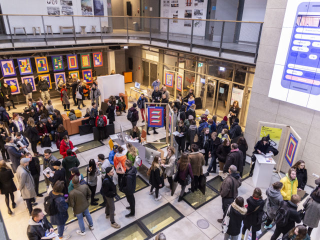 Ludzie zgromadzeni w sali Atrium w budynku B Uniwersytetu Artystycznego w Poznaniu podczas Drzwi Otwartych pomiędzy stoiskami poszczególnych kierunków i wydziałów.