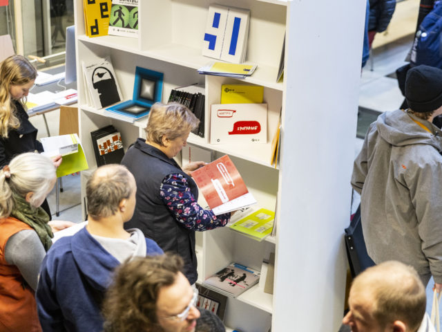 Ludzie zgromadzeni w sali Atrium w budynku B Uniwersytetu Artystycznego w Poznaniu podczas Drzwi Otwartych przeglądający materiały promocyjne oraz książki.
