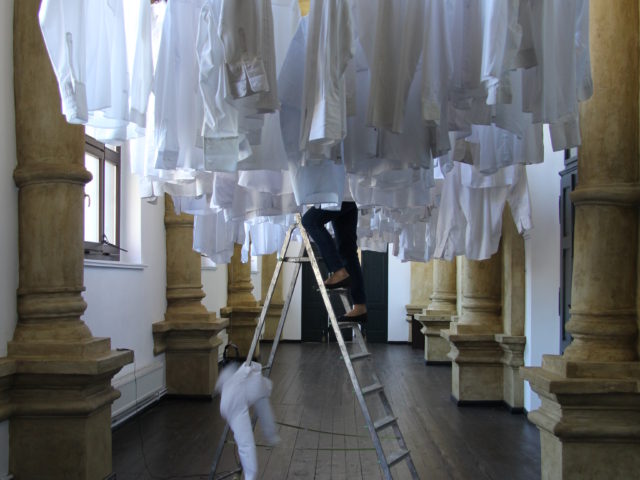 Instalacja artystyczna; wysokie pomieszczenie z kolumnami pośrodku, którego rozwieszone są białe koszule pod sufitem, kobieta zawiesza je stojąc na drabinie.