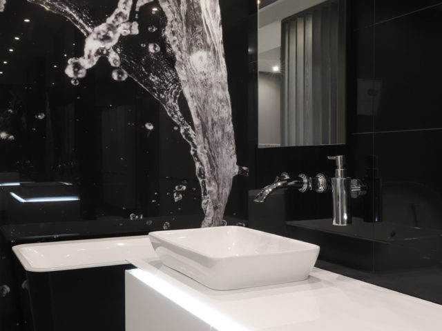 Wnętrze łazienki. Biały sufit, czarne kafelki układające się w powiększenie rozpryśniętej wody. Pod ścianą wanna. Lustro, umywalka i dozownik mydła.