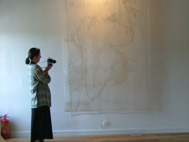 Kobieta z aparatem fotograficznym przed materiałem artystycznym zawieszonym na ścianie. 