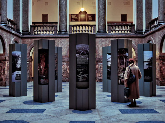 Hol Uniwersytetu Adama Mickiewicza w Poznaniu. Kobieta ogląda wystawę złożoną z metalowych kolumn z obrazami i tekstem.