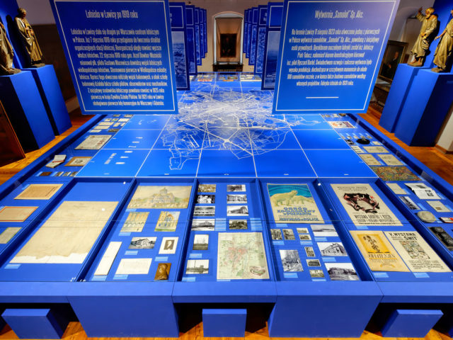 Muzeum Historii Miasta Poznania. Duży niebieski stół; na środku mapa miasta otoczona ekspozycja z pocztówek, przewodników, plakatów map i dokumentów pod szklanym blatem. Podwieszone plansze ze zdjęciami informacją. Po obu stronach pod ścianami stoją figury ludzi.