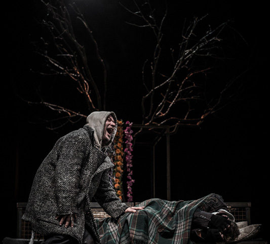 Spektakl teatralny; mężczyzna w płaszczu krzycząc nachyla się i trzyma ręką innego mężczyznę leżącego na ławce owiniętego kocem. W tle drzewo.