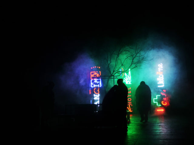 Spektakl teatralny; ludzkie postacie przemieszczające się we mgle oświetlane przez widoczne w tle neony. Zarys drzewa.