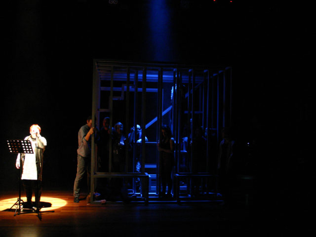 Spektakl teatralny; osoba oświetlona reflektorem przemawia przez mikrofon czytając tekst z pulpitu do nut. Obok duża klatka oświetlona delikatnie niebieskim światłem, w której znajduje się kilka osób.