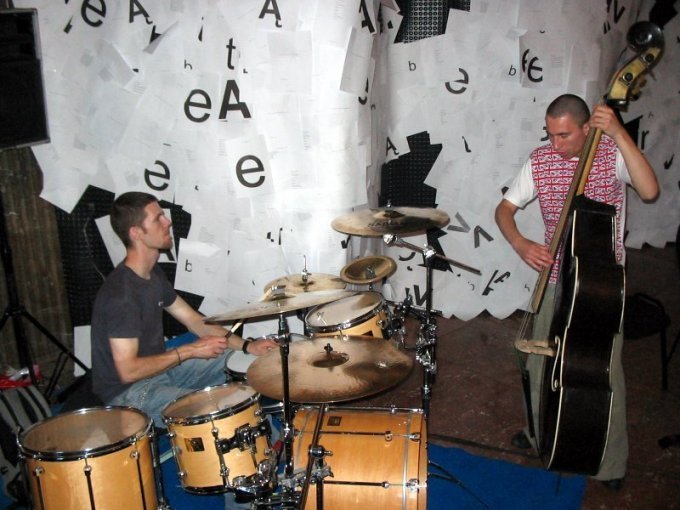 Wnętrze salki muzycznej obklejonej kartkami papieru, gdzie grają perkusista i basista na kontrabasie