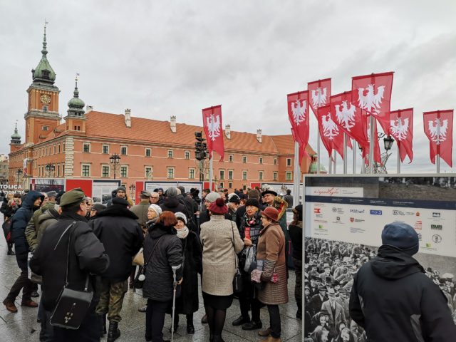 Zgromadzeni ludzie oglądający instalację muzealną na placu Zamkowym w Warszawie z rozstawionych plansz na temat powstania wielkopolskiego.