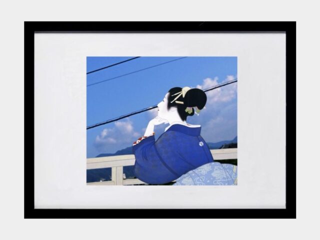 Fotografia ukazująca górzystą, wiejską panoramę Japonii, na kto®ej domalowano kobietę w tradycyjnym stroju japońskim, wpatrującą się z nostalgią w dal opierając się o płot. 