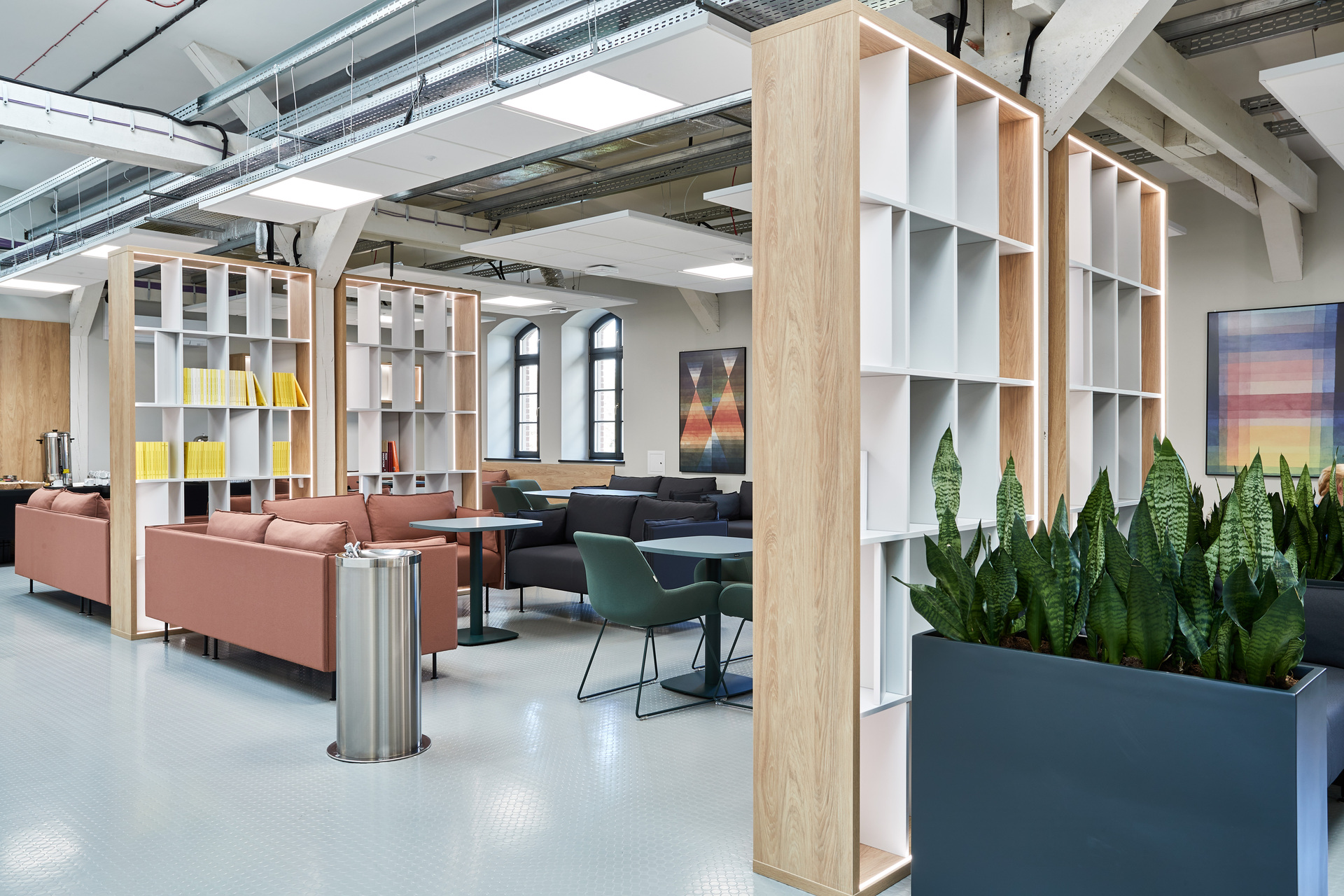 Projekt open space w Uniwersytecie Adama Mickiewicza; przestrzenie z kanapami, fotelami i stolikami oddzielone regałami na książki, śmietniki oraz donice z roślinami; na ścianach obrazy, w tle przestrzeń kuchenna.